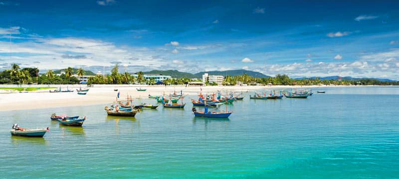 A serene Hua Hin beach with turquoise water, white sand, and small colorful boats anchored near the shore under a clear blue sky.