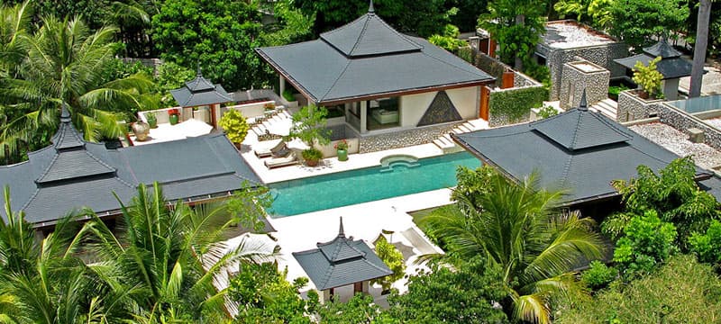 An aerial view of a luxury tropical villa complex in Phuket, Thailand, featuring elegant Thai-style roofs, a long private swimming pool, lush green palm trees, and tranquil garden surroundings.